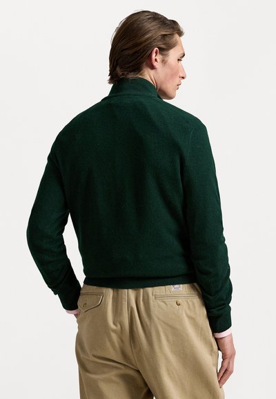 Suéter de gola alta em verde escuro, com bainha e punhos canelados, feito de material suave. Usado com calças cáqui com bolsos traseiros e detalhes em metal.