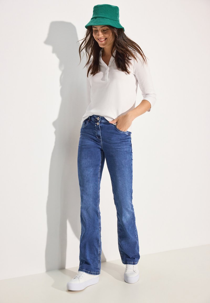 Witte lange mouwen shirt, blauwe uitlopende jeans, groene bucket hat en witte sneakers. Denim textuur met knoopdetails en relaxte pasvorm.