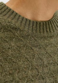 Close-up of a person wearing an olive-green knit sweater with a textured pattern and a ribbed crew neckline.