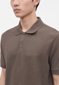 Νέος άνδρας φορώντας καφέ κοντομάνικο πόλο Armani Exchange με γιακά, εμφανίζεται από τον λαιμό έως το μέσο του κορμού σε απλό φόντο.