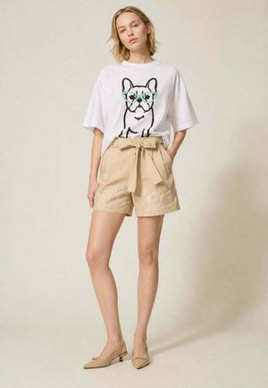 Donna con t-shirt bianca oversize con un cane dei cartoni animati con occhiali, pantaloncini beige a vita alta con fiocco e motivo floreale, e tacchi beige.