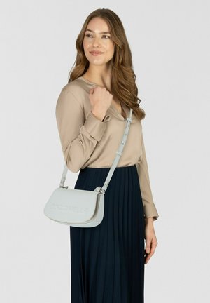 Mujer con blusa beige y falda plisada azul marino que lleva un bolso de hombro Croconelle gris claro con correa ajustable.