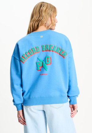 Vrouw met blond haar die een blauwe trui draagt met kleurrijke tekst "Record Breaker PG" en lichtblauwe spijkerbroek, met haar rug naar de camera.