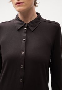 Gros plan d'une personne portant une chemise noire à manches longues boutonnée avec un col pointu sur un fond uni.