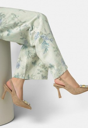 Tacones slingback de ante beige con hebilla con joyas, usados con pantalones florales verde claro, piernas cruzadas mientras está sentado en un taburete cilíndrico blanco.