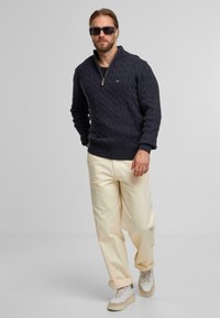 Marineblauwe kabelgebreide trui met een half-rits kraag, gecombineerd met crèmekleurige broeken en lichte sneakers. Eenvoudig ontwerp; geen patronen of accenten.