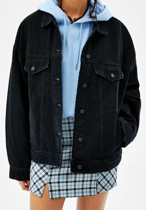 Person trägt eine schwarze Jeansjacke mit Knöpfen über einem hellblauen Hoodie und einem blau-schwarz karierten Minirock mit vorderen Schlitzen.