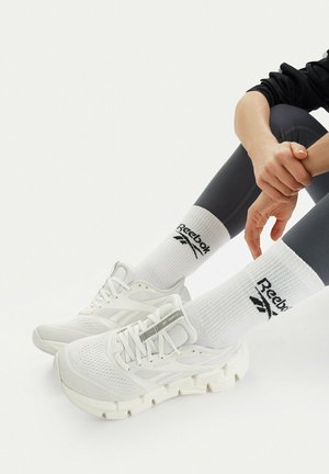 Person wearing white Reebok sneakers and matching white crew socks, sitting with hands resting on bent knee, wearing dark leggings.