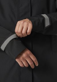 Czarna wodoodporna kurtka z szarymi odblaskowymi akcentami na mankietach. Zbliżenie na dłonie regulujące rękaw, podkreślające materiał i fakturę.