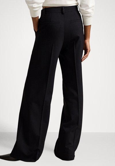 Pessoa vestindo calças pretas de perna larga e uma blusa branca de manga comprida, em pé com as mãos nos bolsos contra um fundo simples.