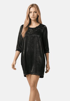 Mujer con cabello largo y rubio que lleva un vestido de terciopelo negro con mangas tres cuartos y encaje floral alrededor del escote, de pie frente a un fondo liso.