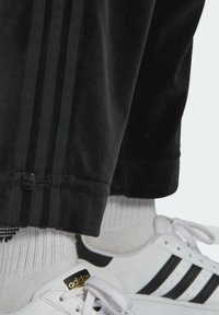 Svarta träningsbyxor har tre vertikala mörka ränder längs sidorna, med en dragsko på mudden. Bärs med vita sneakers.