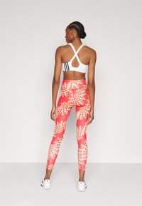 Top desportivo branco com alças cruzadas e riscas pretas, combinado com leggings vermelhas de cintura alta com padrões geométricos de folhas amarelas.