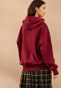 Bordeaux oversized hoodie med hætte, ribbede kanter og dropped shoulders, parret med en ternet plisseret nederdel i mørkegrønne og brune nuancer.