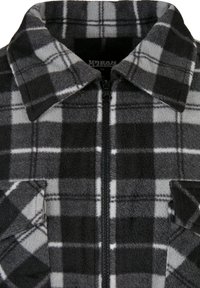 Veste en polaire avec un motif à carreaux noir et gris, dotée d'une fermeture éclair à l'avant, de deux poches poitrine et d'un col pointu. Texture douce.