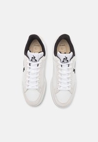 Baskets blanches avec une tige en cuir lisse, des accents en daim gris, un doublage noir, des lacets blancs, et un logo sur la languette et le côté.