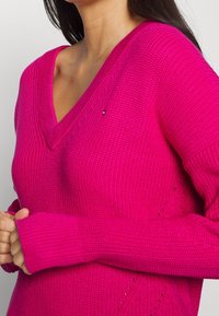 V-Ausschnitt, legerer Strickpullover in leuchtendem Pink. Mit gerippten Bündchen und offenen Details an den Seiten. Weicher Stoff und mittellange Passform.