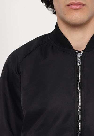 Zwarte bomberjack met een gladde textuur, gereffileerde kraag en een voorrits. Voorzien van gestikte schoudernaden en metallic ritssluiting.