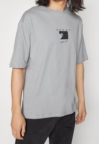 Persona vistiendo una camiseta de gran tamaño en color gris claro con un gráfico negro y el texto "SENSE" en el pecho, combinada con pantalones negros.