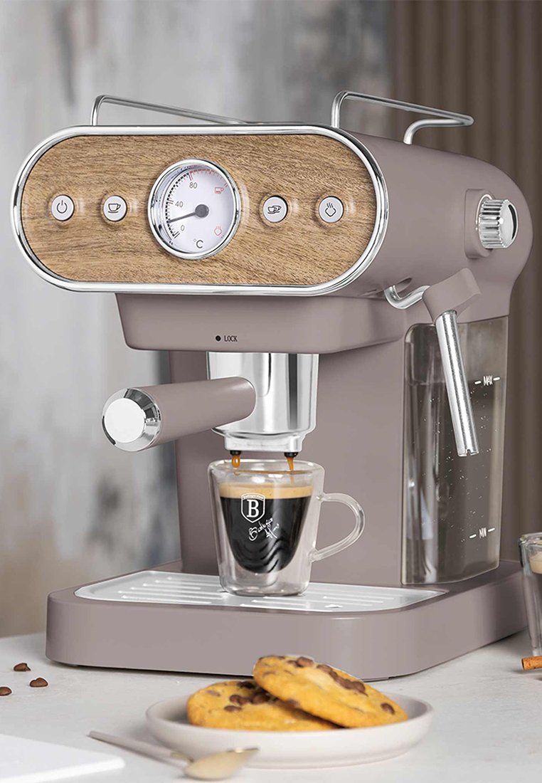 Espresso stroj v jemně růžové barvě s dřevěným akcentem, manometrem a chromovanými knoflíky. Produkuje espresso do průhledného šálku, poblíž jsou čokoládové sušenky s kousky čokolády.