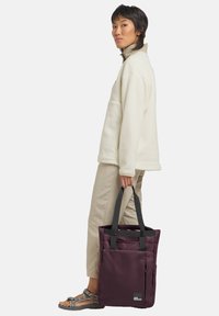 Maroon tote bag with gray straps, zippered pocket, sleek design, made of durable material. Person wearing a cream fleece and beige pants.