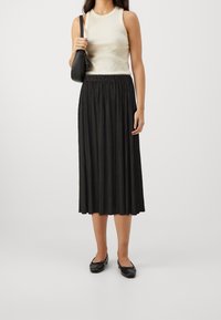 Zwarte geplooide midi-rok met een elastische tailleband, gecombineerd met een beige mouwloos topje en zwarte ballerina's, en een kleine zwarte handtas.