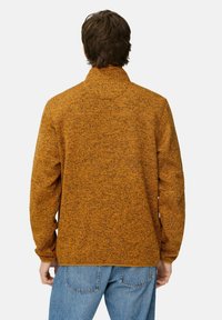 Orange tekstureret jakke med høj krave, med et tofarvet mønster og elastiske manchetter, parret med blå jeans.