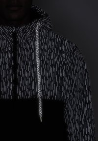 Svart jacka med reflekterande vertikalt randmönster, texturerat tyg, justerbar huva och kontrasterande svart nederdel.