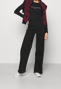 Mustat leveälahkeiset farkut korkealla vyötäröllä, yhdistettynä tiukkaan mustaan puseroon, jossa on valkoinen logo, sekä tummanpunainen huppari, jossa on kuvioitu design.