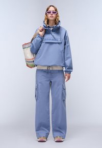 Lys blå, vanntett genser med hette og frontlomme. Matchende cargo-bukser har sidelommer. Modellen bærer en flerfarget totebag.