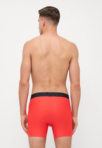 Shorts de boxeur rouges avec une ceinture élastique noire portant le nom de la marque. Le tissu est lisse et ajusté, avec une longueur mi-cuisse.