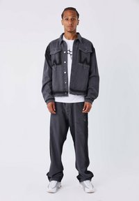 Chaqueta de mezclilla gris oscuro con un diseño cortado, bolsillos frontales y bordes deshilachados, combinada con pantalones holgados a juego y zapatillas blancas.