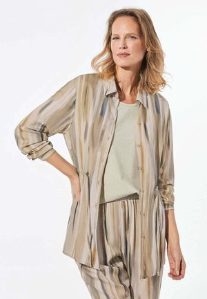 Doorzichtige blouse met lange mouwen en knoopsluiting, uitgevoerd in een beige en bruine gestreepte patroon. Gecombineerd met een lichtgroene top en bijpassende gestreepte broek.