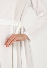 White dress with long sleeves, textured fabric, and a tied waist belt, featuring a pleated skirt and smooth sleeve cuffs.
