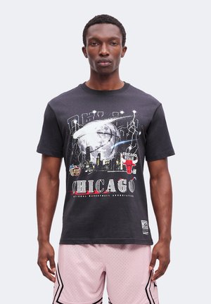 Férfi fekete Chicago Bulls pólóban, melyen kosárlabda, városi sziluett és villám grafikák találhatók, világos rózsaszín kosárlabdás rövidnadrággal párosítva.