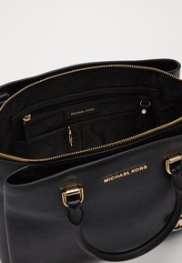 Schwarze Leder-Tote mit goldenen Beschlägen, ausgestattet mit einem Reißverschlussverschluss und Innenfächern, geprägtem Logo und strukturiertem Äußeren.