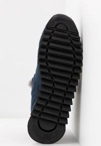 Semelle de chaussure en caoutchouc noir avec des rainures profondes et larges conçues pour la traction, affichée verticalement sur un fond blanc avec angles.