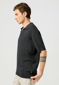 Kortärmad polo i mörkgrått, med stickad textur, spetsig krage och sidofickor för ökad rörelsefrihet.