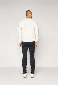 Dugih rukava off-white majica s okruglim izrezom i detaljem na leđima, uparen s crnim uskim hlačama i svijetlim tenisicama.