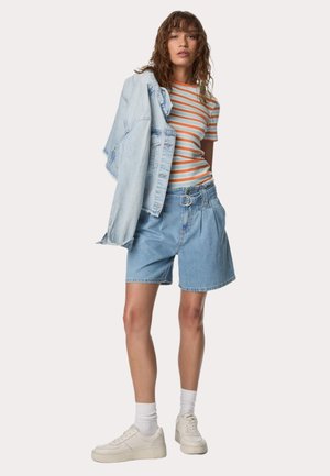 Denimshorts och randig t-shirt i orange och vitt. Ljusblå denimjacka hängande över axeln. Vita sneakers och strumpor som når anklarna.