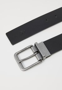 Cintura in pelle nera con una fibbia metallica rettangolare argentata con branding visibile. Il cinturino presenta cinque fori e una texture liscia.