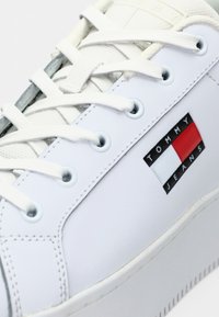 White leather sneaker with flat laces, a gray accent, and a rectangular TOMMY JEANS logo featuring red, white, and blue colors on the side.