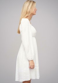 Vestido blanco de longitud a la rodilla con una tela texturizada, mangas abullonadas y un corpiño ajustado. Silueta suave con parte inferior en capas y un patrón sutil.