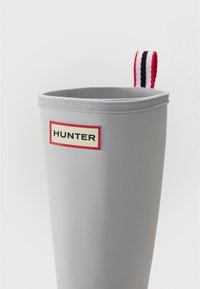 Grå gummistävla med rundad topp, med en röd och krämfärgad "HUNTER"-logotyp och en flerfärgad randig tygögla.