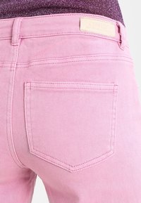 Jeans de mezclilla rosa con un diseño ajustado, que cuentan con bolsillos traseros y detalles de costura sutil. La etiqueta adjunta muestra el nombre de la marca.