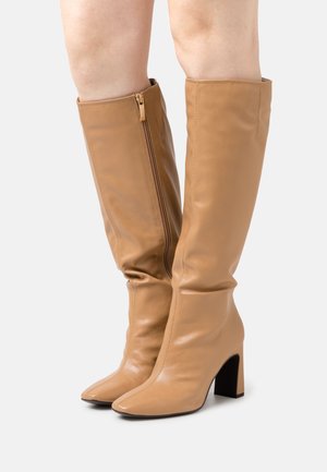 Bottes beiges montantes en cuir lisse avec une fermeture éclair sur le côté, bout pointu et talon épais. Présente un design décontracté pour un confort supplémentaire.