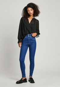 Μαύρη, διάφανη μπλούζα με υφασμάτινες κουκίδες και ριχτές μανίκες, σε συνδυασμό με ψηλοτάκουνες μπλε τζιν παντελόνες και γυαλιστερές μαύρες λιτές εσπαντρίγιες.