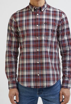 Homme portant une chemise à carreaux à manches longues rouge, blanche et bleue avec une poche poitrine et un jean bleu.
