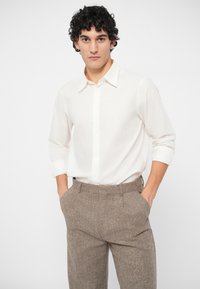 Mann med krøllete, mørkt hår har på seg en hvit skjorte med lange ermer som er stukket inn i teksturerte brune bukser, står med hendene i lommene mot en hvit bakgrunn.