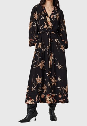 Kobieta w czarnej midi sukience kopertowej z brązowymi kwiatowymi wzorami, długimi rękawami, kieszeniami oraz czarnych szpilkach z ostrym noskiem.